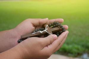 Wooden cross and wooden rosary are held in hands of young asian Catholic prayer while praying in the temple park area. photo