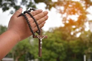 la cruz de madera y el rosario de madera se sostienen en manos de jóvenes católicos asiáticos rezando mientras rezan en el área del parque del templo. foto