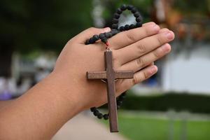 la cruz de madera y el rosario de madera se sostienen en manos de jóvenes católicos asiáticos rezando mientras rezan en el área del parque del templo. foto