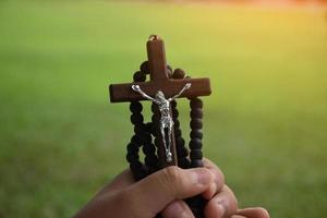la cruz de madera y el rosario de madera se sostienen en manos de jóvenes católicos asiáticos rezando mientras rezan en el área del parque del templo. foto