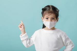 Asian little child girl wearing respirator mask to protect coronavirus outbreak and pointing hand to blank background, New virus Covid-19 from Wuhan China concept, Empty space isolated on blue photo