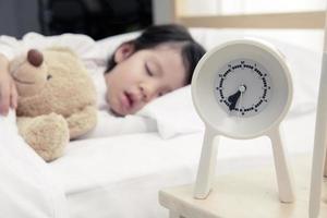 Child girl sleeping with alarm clock on the wooden bed in her bedroom, Happy asian child little girl and alarm clock wake up in the morning photo