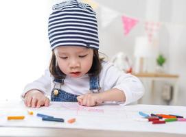 Little asian girl sitting at table in room, Preschooler girl drawing on paper with colorful pens on sunny day, kindergarten or photo