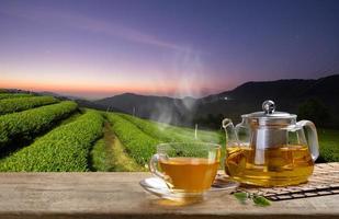 Cup of hot tea and glass jugs or jars and tea leaf on the wooden table and the tea plantations background photo