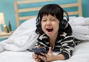 una niña linda canta una canción con un smartphone en su dormitorio, una niña asiática feliz escuchando música con auriculares en la cama, concepto de tecnología foto