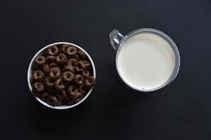 un vaso de leche sobre un fondo negro y anillos de maíz con chocolate. delicioso desayuno de copos de maíz y leche. foto