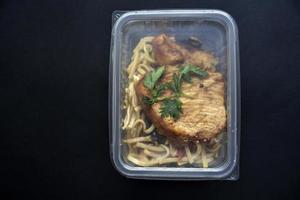 A piece of juicy fried pork with seasoned noodles in a container. Delicious breakfast with meat and noodles on a black background. photo
