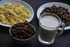 A saucer and a cup of cornflakes and a glass of milk on a black background. Delicious breakfast of cereal with honey and chocolate with milk. photo