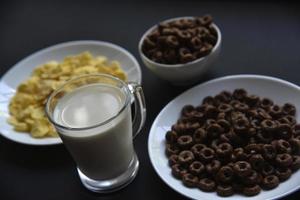 A saucer and a cup of cornflakes and a glass of milk on a black background. Delicious breakfast of cereal with honey and chocolate with milk. photo