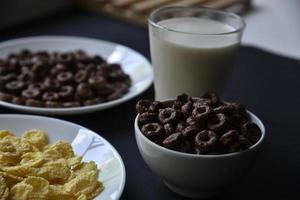 un platillo y una taza de copos de maíz y un vaso de leche sobre un fondo negro. delicioso desayuno de cereal con miel y chocolate con leche. foto