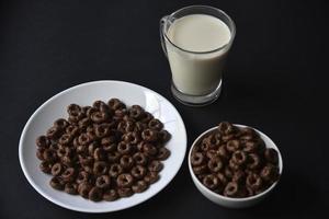 A saucer and a cup of cornflakes and a glass of milk on a black background. Delicious breakfast of cereal with honey and chocolate with milk. photo