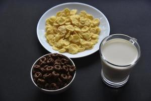 A saucer and a cup of cornflakes and a glass of milk on a black background. Delicious breakfast of cereal with honey and chocolate with milk. photo