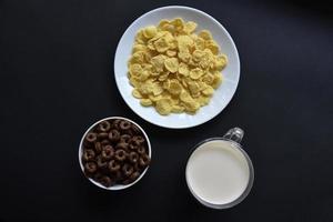 un platillo y una taza de copos de maíz y un vaso de leche sobre un fondo negro. delicioso desayuno de cereal con miel y chocolate con leche. foto