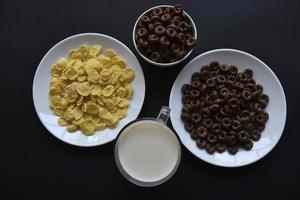 un platillo y una taza de copos de maíz y un vaso de leche sobre un fondo negro. delicioso desayuno de cereal con miel y chocolate con leche. foto