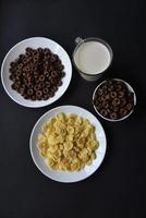 A saucer and a cup of cornflakes and a glass of milk on a black background. Delicious breakfast of cereal with honey and chocolate with milk. photo