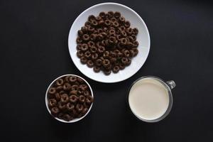 un platillo y una taza de copos de maíz y un vaso de leche sobre un fondo negro. delicioso desayuno de cereal con miel y chocolate con leche. foto