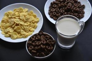 A saucer and a cup of cornflakes and a glass of milk on a black background. Delicious breakfast of cereal with honey and chocolate with milk. photo