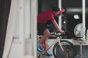 hombre en bicicleta en el entrenador de la máquina que está haciendo ejercicio en el hogar por la noche jugando al juego de carreras de bicicletas en línea foto