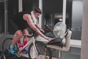 hombre en bicicleta en el entrenador de la máquina que está haciendo ejercicio en el hogar por la noche jugando al juego de carreras de bicicletas en línea foto