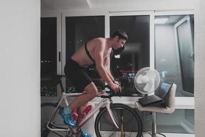 hombre en bicicleta en el entrenador de la máquina que está haciendo ejercicio en el hogar por la noche jugando al juego de carreras de bicicletas en línea foto