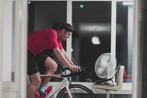 hombre en bicicleta en el entrenador de la máquina que está haciendo ejercicio en el hogar por la noche jugando al juego de carreras de bicicletas en línea foto