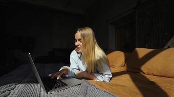 Young blonde woman lays on couch in sunlight from window uses laptop video