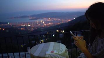 mulher se senta à mesa da janela usando telefone inteligente com vista para uma cidade à noite video