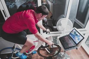 hombre en bicicleta en el entrenador de la máquina que está haciendo ejercicio en el hogar por la noche jugando al juego de carreras de bicicletas en línea foto