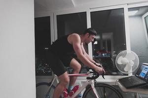 hombre en bicicleta en el entrenador de la máquina que está haciendo ejercicio en el hogar por la noche jugando al juego de carreras de bicicletas en línea foto