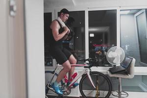 hombre en bicicleta en el entrenador de la máquina que está haciendo ejercicio en el hogar por la noche jugando al juego de carreras de bicicletas en línea foto