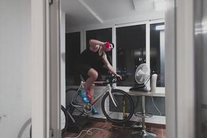 hombre en bicicleta en el entrenador de la máquina que está haciendo ejercicio en el hogar por la noche jugando al juego de carreras de bicicletas en línea foto