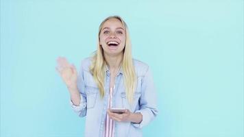 Young blonde woman in front of light blue backdrop smiles and waves video