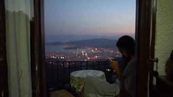 Woman sits at window table using smart phone overlooking a city at night video