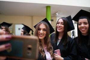 group of happy international students in mortar boards and bachelor gowns with diplomas taking selfie by smartphone photo