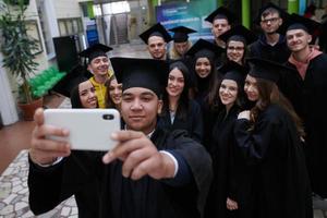 group of happy international students in mortar boards and bachelor gowns with diplomas taking selfie by smartphone photo