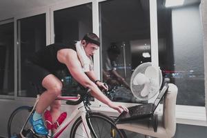 hombre en bicicleta en el entrenador de la máquina que está haciendo ejercicio en el hogar por la noche jugando al juego de carreras de bicicletas en línea foto