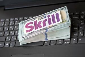 TERNOPIL, UKRAINE - SEPTEMBER 6, 2022 Skrill paper logotype lies on black laptop with US dollar bills. Payoneer is American financial services company provides online money transfer photo