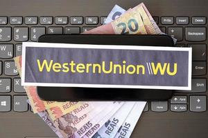 TERNOPIL, UKRAINE - SEPTEMBER 6, 2022 Western Union paper logotype lies on black laptop with brazilian reals bills. Western Union Company is American multinational financial services company photo