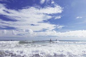 turistas divirtiéndose, disfrutando del surf y nadando alegremente en el mar tropical con olas, cielo azul y nubes blancas en el fondo, hermosa vista agradable día soleado. foto