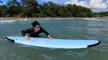 Young boy, student in surfing, is holding onto softboard and trying to bring it back into sea to practice while playing against waves and splashing water. photo