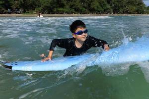 Young boy, student in surfing, holding onto softboard and trying to bring it back into sea to practice while playing against waves and splashing water. photo