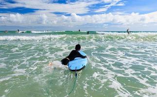 Young boy, a beginner in surfing, holding softboard and trying to bring it back into sea to practice while playing against waves and splashing water. photo