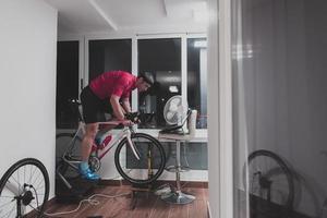 hombre en bicicleta en el entrenador de la máquina que está haciendo ejercicio en el hogar por la noche jugando al juego de carreras de bicicletas en línea foto