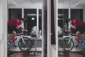 hombre en bicicleta en el entrenador de la máquina que está haciendo ejercicio en el hogar por la noche jugando al juego de carreras de bicicletas en línea foto