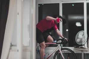 hombre en bicicleta en el entrenador de la máquina que está haciendo ejercicio en el hogar por la noche jugando al juego de carreras de bicicletas en línea foto