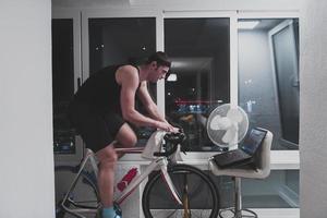 hombre en bicicleta en el entrenador de la máquina que está haciendo ejercicio en el hogar por la noche jugando al juego de carreras de bicicletas en línea foto