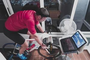 hombre en bicicleta en el entrenador de la máquina que está haciendo ejercicio en el hogar por la noche jugando al juego de carreras de bicicletas en línea foto