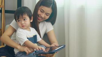 madre e hijo asiáticos se sientan en casa y trabajan con tablats en el trabajo. mamá trabajando en línea hijo sentado en su regazo video