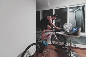 hombre en bicicleta en el entrenador de la máquina que está haciendo ejercicio en el hogar por la noche jugando al juego de carreras de bicicletas en línea foto