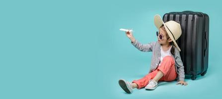 Asian little girl tourist woman playing airplane model toy with black luggage to travel on weekends, empty space in studio shot isolated on colorful blue long banner background photo